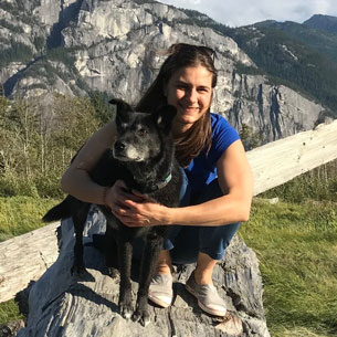 Dr. Tanya Wright, DVM, DACVS-SA, smiling and crouching beside her black dog with towering mountains and green trees in the background on a sunny day.