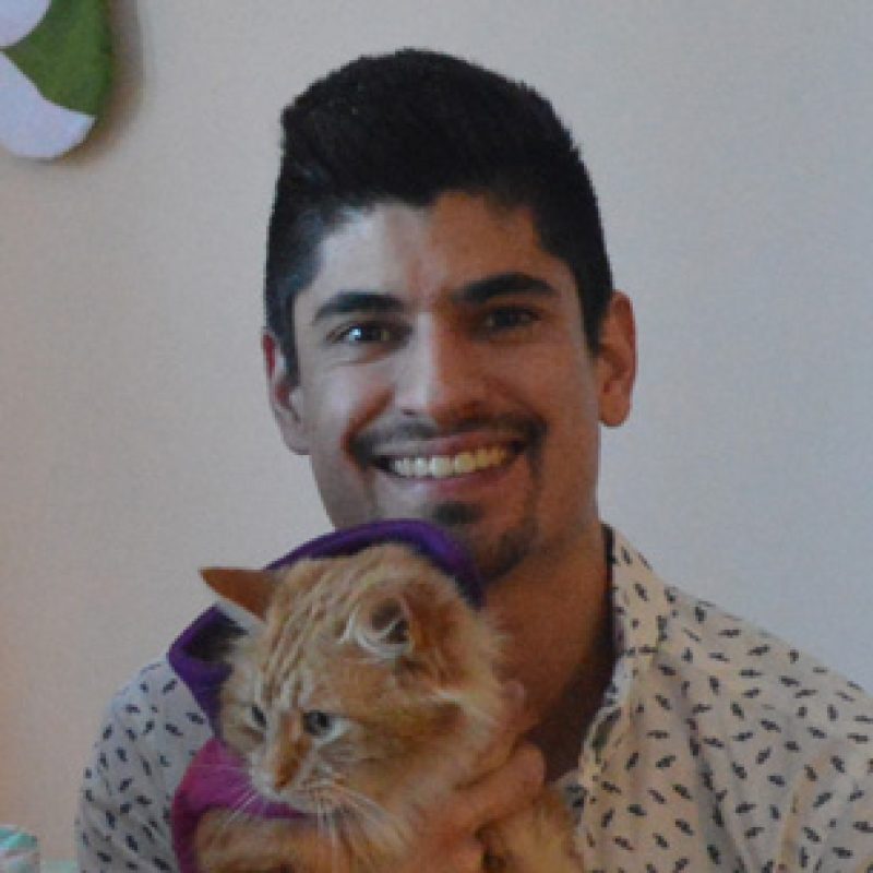 Dr. Alex Alvarez, Ldo Vet, MS, Dipl. ACVS-SA, smiling while holding an orange cat indoors, with a light-colored wall in the background.