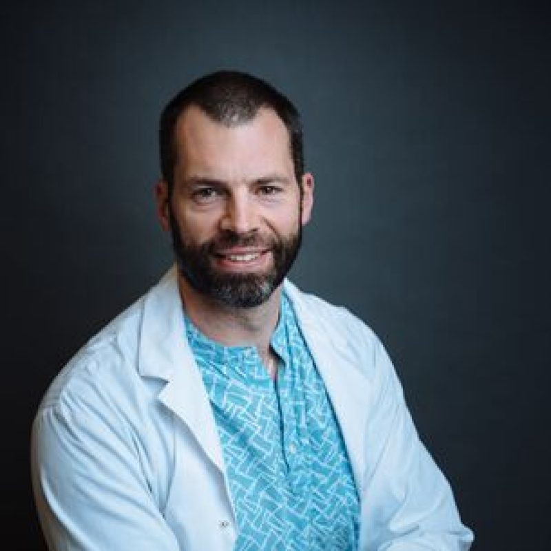 Dr. Evan Crawford, DVM, MSc, DVSc, DACVS-SA, smiling while wearing a white lab coat over a blue patterned shirt, posed against a dark background.