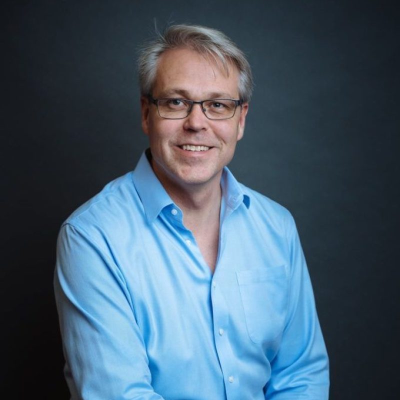 Dr. Geoffrey Hutchinson, DVM, MS, DACVS, smiling while wearing a light blue dress shirt and glasses, posed against a dark background.
