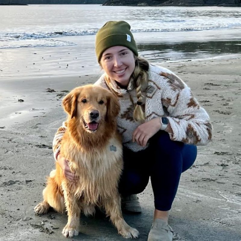 Dr. Jovana Boljanovic, Emergency Veterinarian at Boundary Bay Veterinary Specialty Hospital, kneeling on a beach beside a Golden Retriever